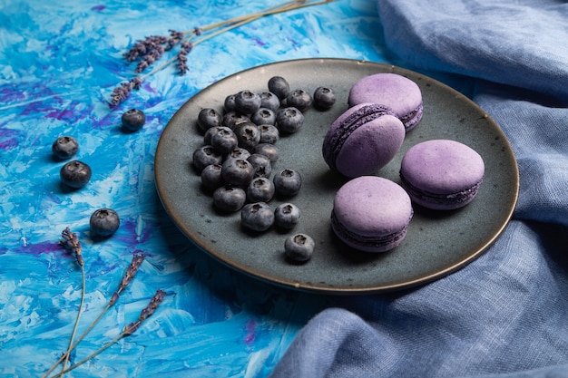 Purple macarons or macaroons cakes with blueberries on ceramic plate on a blue concrete surface and blue textile