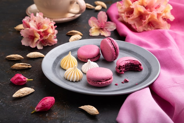 Purple macarons or macaroons cakes with cup of coffee on a black concrete background and pink textile. Side view,