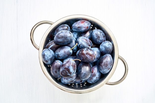 Purple plums in colander on white wooden table. 