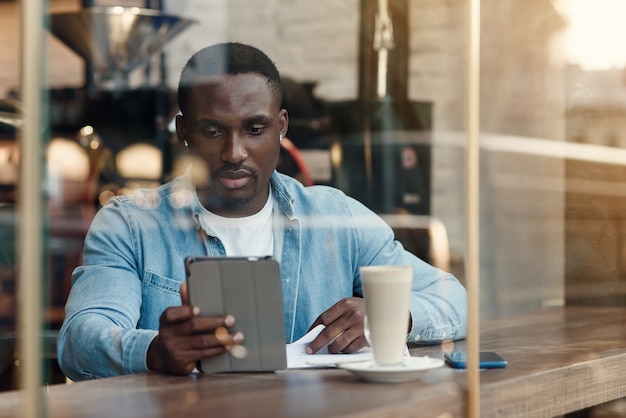 Purposeful black male entrepreneur uses tablet pc while sitting in cafe with coffee near window