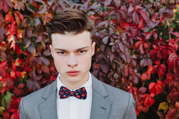 Purposeful young guy posing in a suit with a bow tie on the\
background of a beautiful bush with bright red leaves.