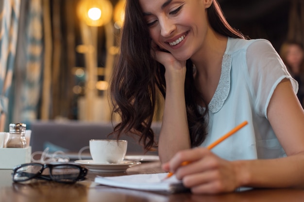 Put it down. Cheerful merry thoughtful woman grinning while writing down plan and drinking coffee