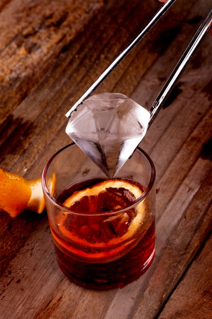 Putting diamond ice into red negroni drink with campari and vermouth on wooden table with orange top closeup