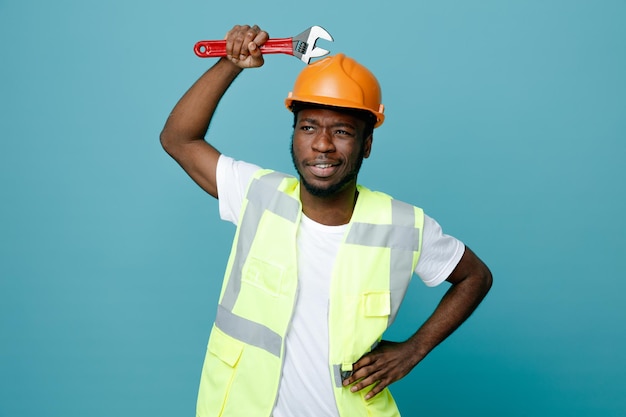 Putting hand on hips young african american builder in uniform holding gas wrench on head isolated on blue background