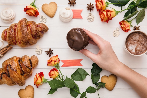Выложить домашний шоколадный кекс на белый украшенный деревянный стол с круассанами и цветами на День рождения, День святого Валентина.