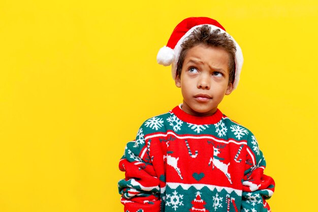 Puzzled african american boy in christmas clothes thinks and plans on yellow isolated background