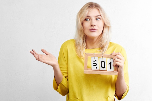 Puzzled attractive woman in yellow clothes with calendar thinks about preparing for vacation trip, not much time left until first day of summer