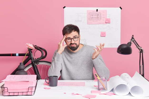Puzzled bearded man raises hand keeps hand on temple faces difficult task cannot find solution wears casual clothes poses in coworking space against pink wall. Hesitant male architect.