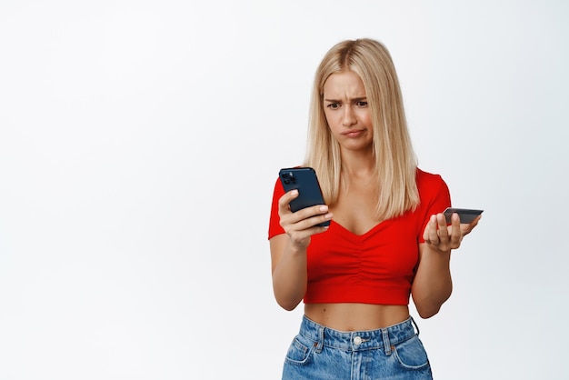 Puzzled blond woman stares at her phone while shopping online holds credit card stands against white background