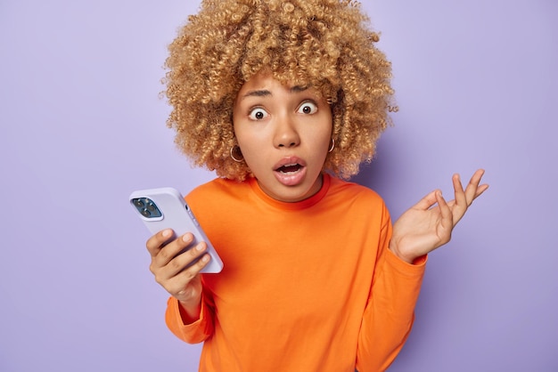 Puzzled confused woman shrugs shoulders looks unaware stares\
with omg expression holds mobile phone checks notification wears\
orange jumper isolated over purple background human reactions\
concept