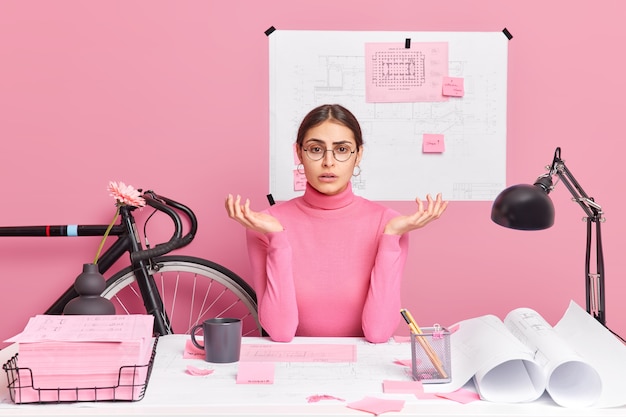 Puzzled disappointed European woman office worker spreads palms creats graphic feels confused wears round spectacles turtleneck poses in coworking space has blueprints and sketches on desktop