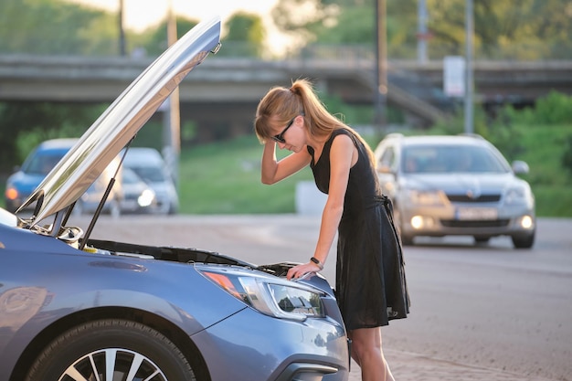 Puzzled female driver standing on a city street near her car with popped up hood looking at broken engine