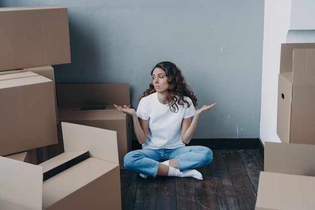 Puzzled girl shrugging sitting with boxes Hard relocation day to new home Moving company service