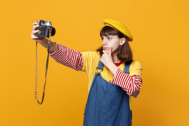 Adolescente perplessa in berretto francese che fa selfie girato su una fotocamera vintage retrò, mettendo la mano sul mento isolato su sfondo giallo. emozioni delle persone, concetto di stile di vita. mock up spazio di copia.