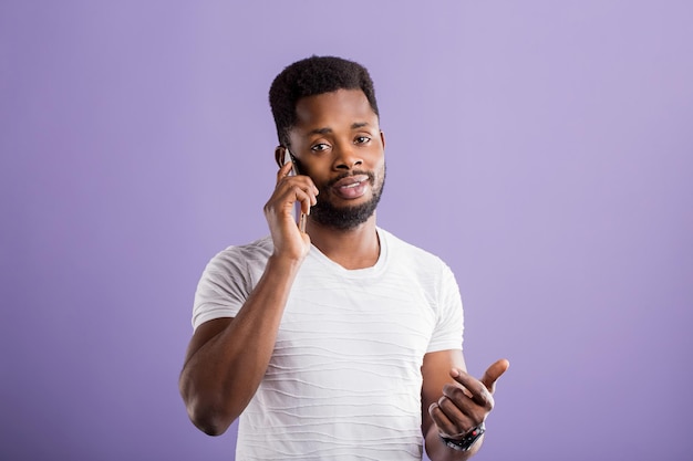 어리둥절한 수염을 기른 아프리카 남성이 스마트폰으로 이야기하고, 카메라를 혼란스럽게 바라보고, 보라색 배경 위에 손으로 서 있는 몸짓을 합니다. 얼굴 표정과 반응 개념.