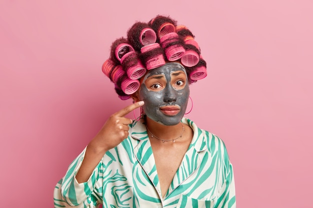 Puzzled housewife undergoes beauty procedures has problematic skin points at clay mask on face reduces fine lines applies rollers for making hairstyle wears dressing gown isolated over pink wall