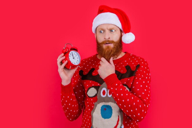 Puzzled man in Santa Claus hat stroking beard looking at alarm clock time New Year countdown