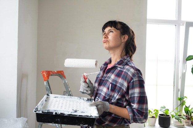 Puzzled woman in paint roller and white paint for walls in hands closeup portrait Construction work and cosmetic repairs in house wall painting tinting finishing work with your own hands