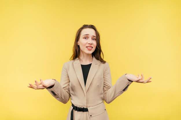 Puzzled woman in smart casual clothes stands on a yellow background and spreads her arms away