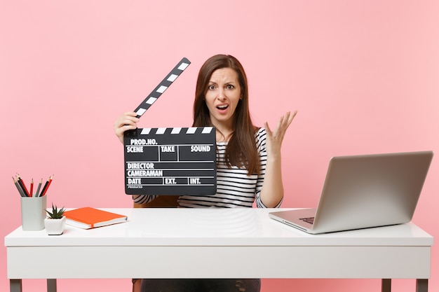 Puzzled woman spreading hands holding classic black film making clapperboard, working on project while sit at office with laptop