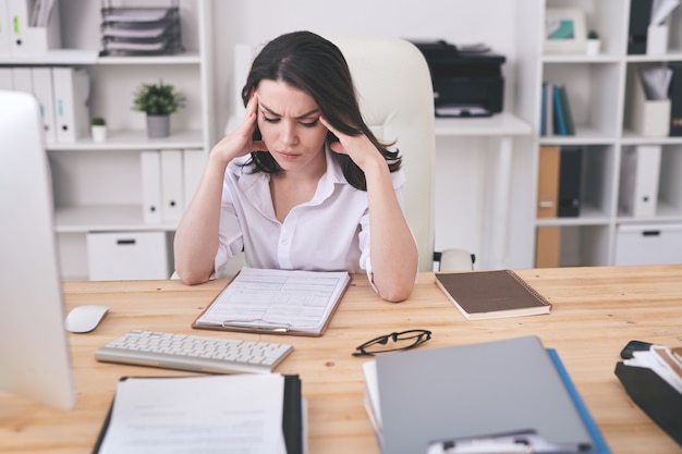 Puzzled young businesswoman sitting at desk in modern office and analyzing contract content