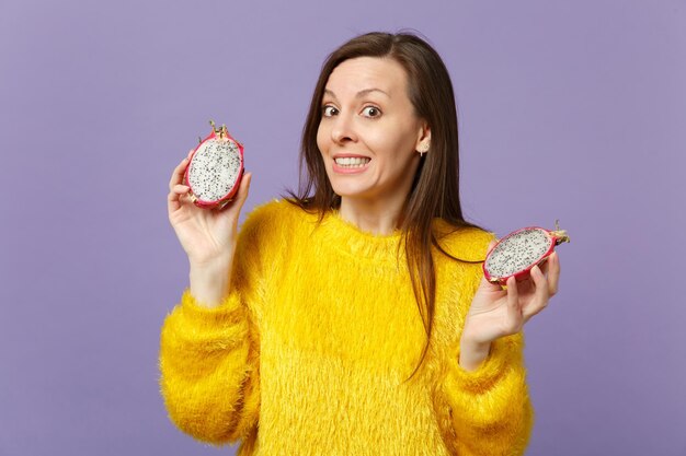 Puzzled young woman in fur sweater holding in hands halfs of fresh ripe pitahaya, dragon fruit isolated on violet pastel background. People vivid lifestyle relax vacation concept. Mock up copy space.