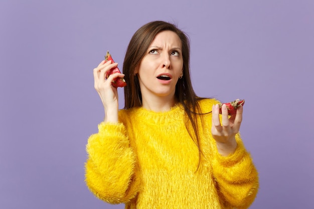 Puzzled young woman in fur sweater looking up holding halfs of pitahaya, dragon fruit near ears isolated on violet pastel background. People vivid lifestyle relax vacation concept. Mock up copy space.