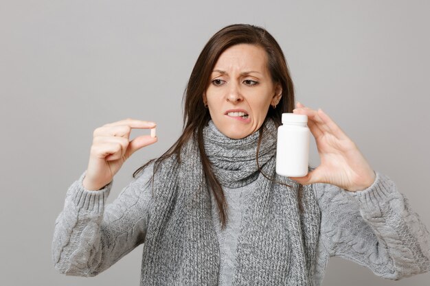 Giovane donna perplessa in maglione grigio, sciarpa che morde le labbra che tengono compresse di farmaci pillole di aspirina in bottiglia isolato su sfondo grigio. stile di vita sano malato malato trattamento stagione fredda concetto.