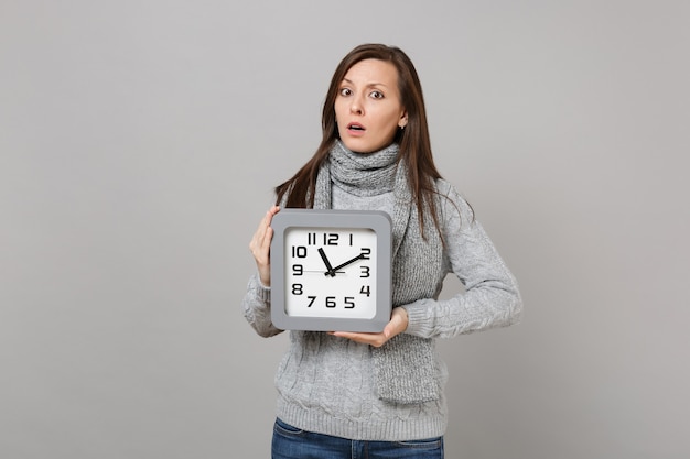 Puzzled young woman in gray sweater, scarf hold square clock isolated on grey background, studio portrait. Healthy fashion lifestyle, people sincere emotions, cold season concept. Mock up copy space.