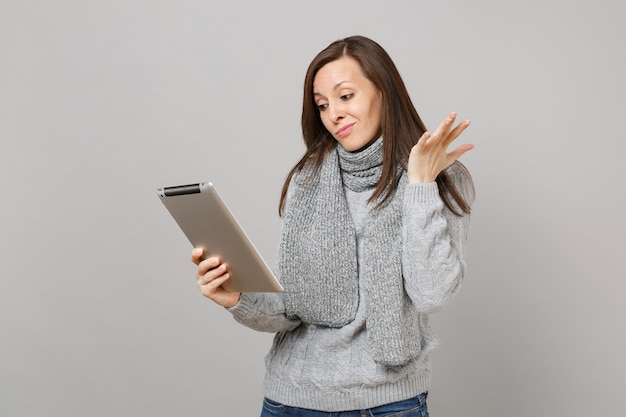 Puzzled young woman in gray sweater, scarf spreading hand using tablet pc computer isolated on grey background. Healthy lifestyle, online treatment consulting, cold season concept. Mock up copy space.