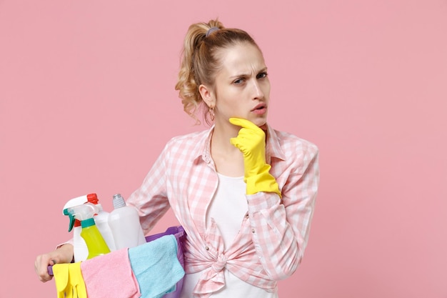 Puzzled young woman housewife in rubber gloves hold basin with detergent bottles washing cleansers doing housework isolated on pink background studio. Housekeeping concept. Put hand prop up on chin.