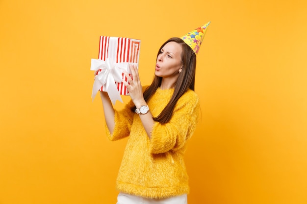 明るい黄色の背景で隔離の休日を祝って楽しんでいるギフトプレゼントと赤い箱に何が入っているかを推測しようとしている誕生日パーティーハットの困惑した若い女性。人々は誠実な感情、ライフスタイル。