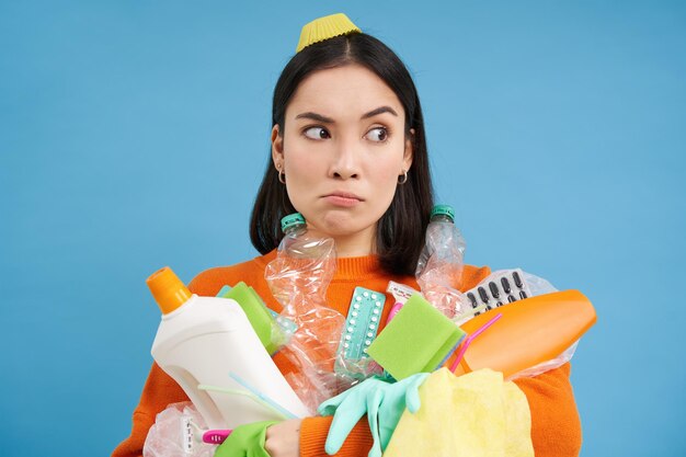 Puzzled young woman with pile of garbage holds plastic bottles and trash for recycling looks perplex