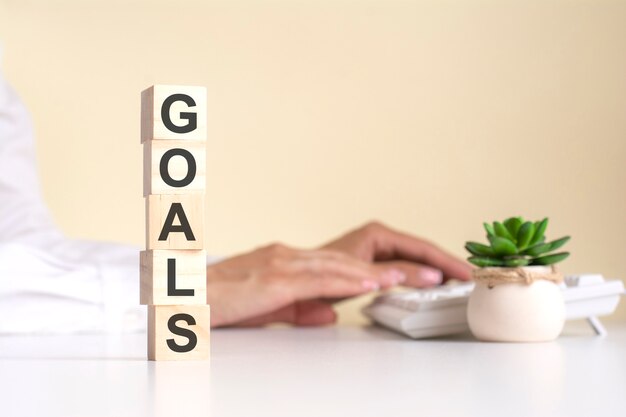 Pyramid of wooden cubes with the word goals in the foreground, in the background - business woman works on the keyboard in a light office, free space. selective focus