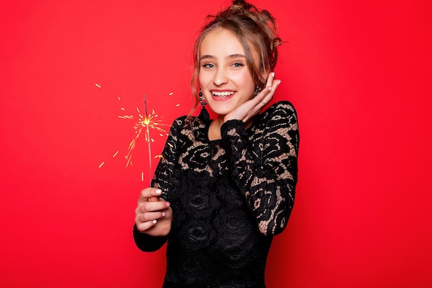 Pyrotechnics and people concept - smiling young woman or teenage girl happy woman with sparklers celebrate in black dress on red background. girl is surprised