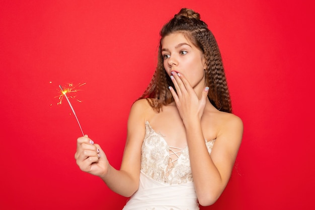 Pyrotechnics and people concept - smiling young woman or teenage girl happy woman with sparklers celebrate in white dress on red background. surprised with his hand and look up