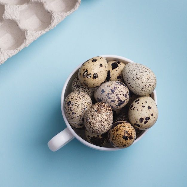 Quail eggs in cup and cardboard packaging on blue background.