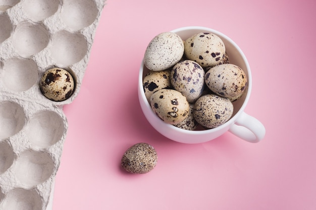 Quail eggs in cup and cardboard packaging on pink background.