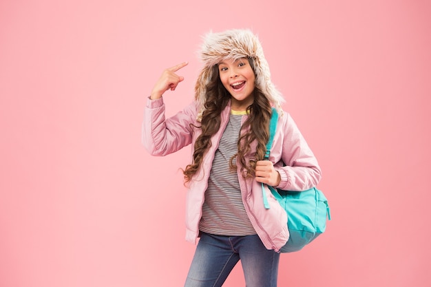 Quality School. small happy girl back to school. winter holiday vacation. kid warm clothes pink background. happy child earflap hat. schoolgirl with bag go home. end of school year. no more exams.