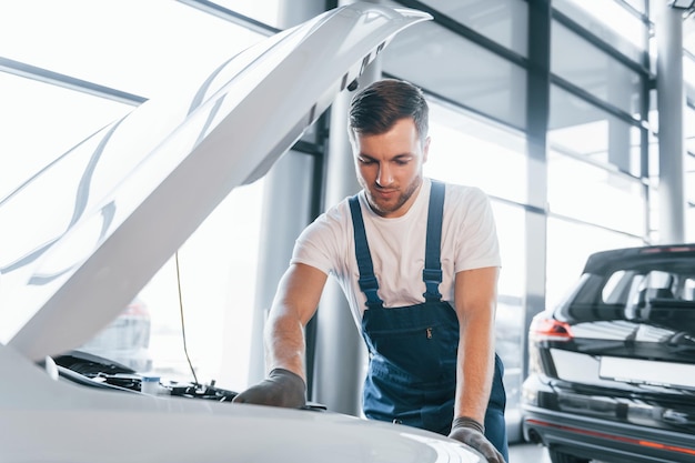 Качественное обслуживание Молодой человек в белой рубашке и синей форме ремонтирует автомобиль