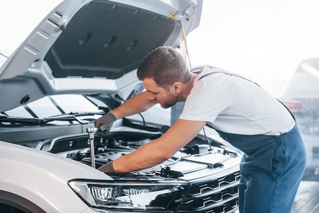 Quality service Young man in white shirt and blue uniform repairs automobile