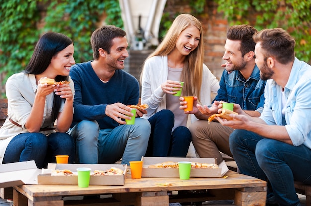 Quality time with friends. Group of joyful young people talking to each other and eating pizza while sitting outdoors