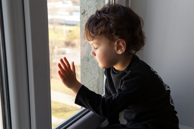 Quarantine during a coronavirus pandemic. . The child stands on the windowsill and looks at the empty city through the window. Dreams to go out and breathe fresh air.