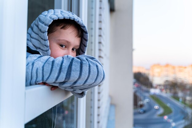 Quarantine Little boy stands and look out of a window bored Longing for fresh air and walks along the street Forced home during quarantine due to the coronavirus pandemic