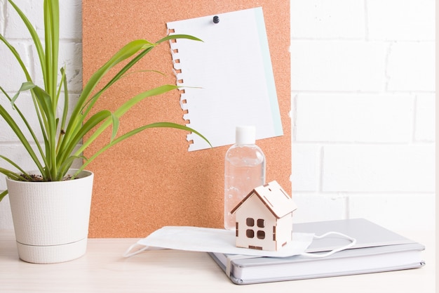 Photo quarantine workplace at home with face mask and gel sanitizer, copy space, potted plant, cork board and notepad on the table