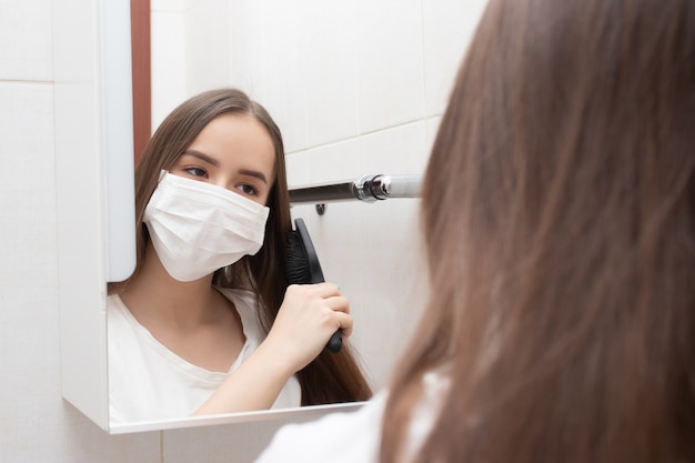 Quarantined life. A woman in a medical mask looks in the mirror, combing her hair. Prevention and protection against diseases