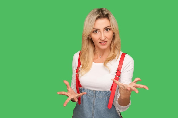 Quarrel aggressive emotions Portrait of irritated adult blond woman in stylish denim overalls raising her hands in anger clenching teeth and looking furious annoyed indoor studio shot isolated