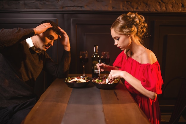 Quarrel of young couple in restaurant, bad evening. Elegant woman in red dress and her man eating in cafe