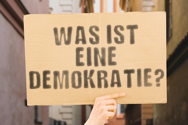 The question What is a democracy is on a banner in men's hands with blurred background Tolerance Debate Compromise Negotiation Coalition Awareness Education Advocacy Campaign