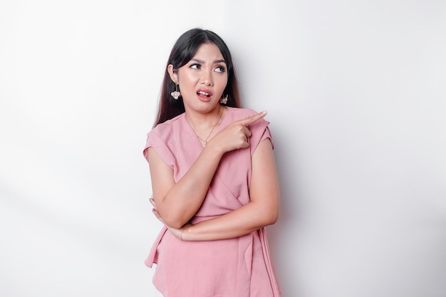 그녀의 표정 옆에있는 젊은 아시아 인 포인팅 복사 공간 질문 혼란스러운 입을 벌리고 불확실한 우둔한 스탠드 흰색 배경 착용 분홍색 블라우스